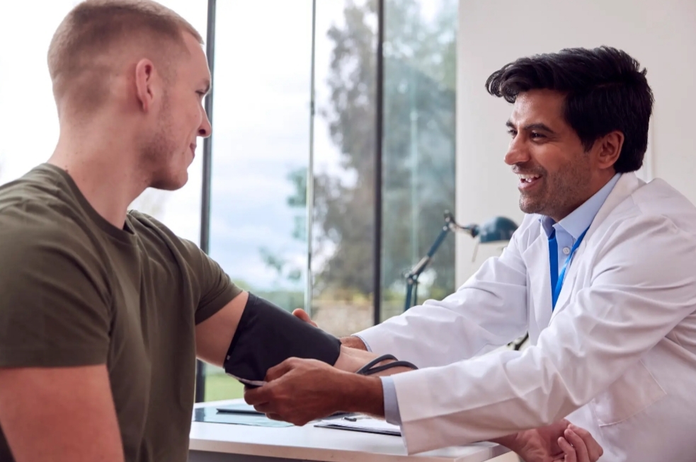 A doctor checks a man's blood pressure during a routine health examination in a modern clinic. The patient is smiling while wearing a blood pressure cuff, highlighting the importance of regular preventive health checkups.